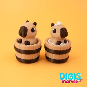 May include: Two tan and black capybara figurines in wooden tubs. The capybaras have black ears and noses, with black spots on their bodies. One has white foam around it. The tubs have dark brown bands. The background is yellow.