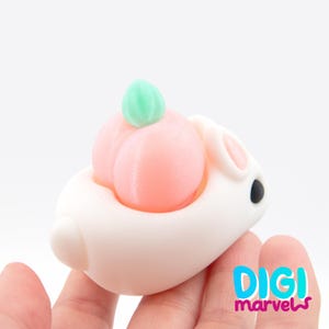May include: A white bunny-shaped figurine with a peach on its back. The peach is light pink with a green leaf on top. The bunny has black eyes and pink inner ears. The Digi Marvels logo is in the bottom right corner.