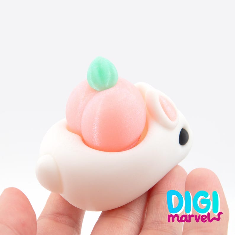 May include: A white bunny-shaped figurine with a peach on its back. The peach is light pink with a green leaf on top. The bunny has black eyes and pink inner ears. The Digi Marvels logo is in the bottom right corner.