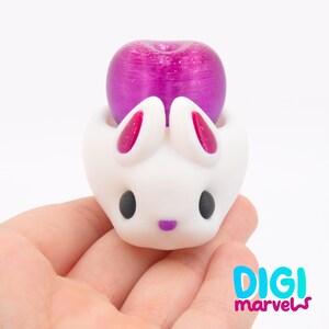 May include: A white bunny-shaped figurine with a purple fruit on top. The bunny has black eyes, pink inner ears, and a purple nose. The figurine is held in a hand.