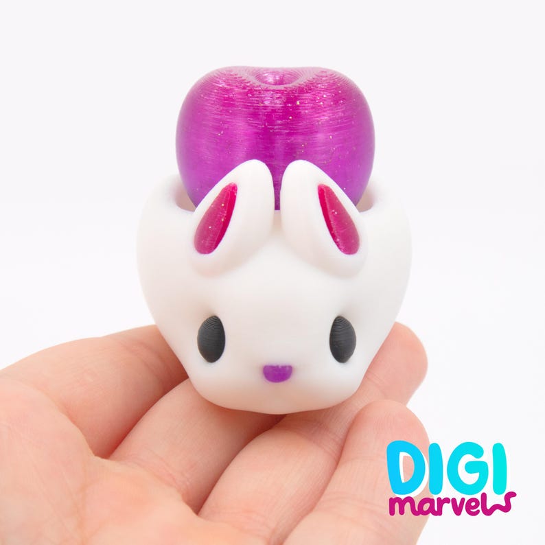 May include: A white bunny-shaped figurine with a purple fruit on top. The bunny has black eyes, pink inner ears, and a purple nose. The figurine is held in a hand.