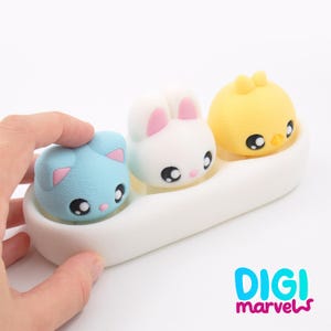 May include: A collection of three cute animal figurines: a blue cat, a white rabbit, and a yellow chick, each with big, round eyes. They are presented in a white, curved container. The Digi Marvels logo is visible in the bottom right corner.