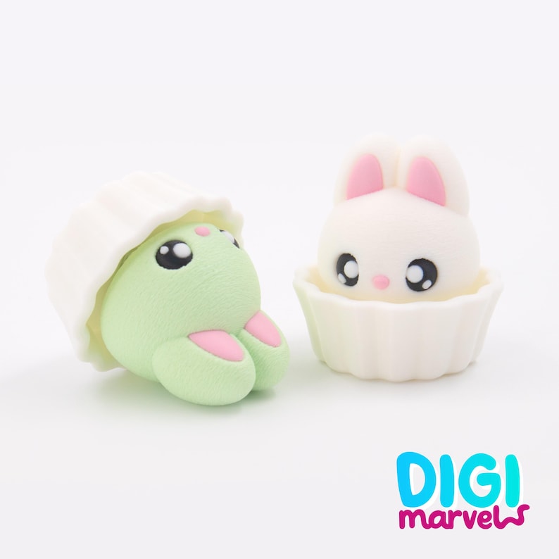 May include: Two cute figurines: a green creature with pink details and a white bunny with pink ears, both in white cupcake holders. They have large black eyes and are on a white background. The Digi Marvels logo is visible.