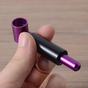 May include: A purple and black metal smoking pipe with a removable bowl. The pipe is held in a hand.