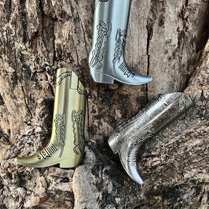 Puede incluir: Tres figuras en miniatura de botas de vaquero en tonos dorados, plateados y bronce. Cada bota presenta costuras decorativas y una punta puntiaguda. Las botas están dispuestas sobre una superficie marrón texturizada, lo que sugiere un tema rústico.