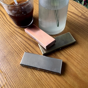May include: Three metal cigarette cases, one with a textured silver finish, one with a copper finish, and one with a brass finish. The cases are on a wooden surface.