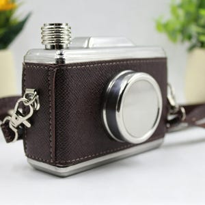May include: A brown leather-wrapped flask shaped like a vintage camera. It has a silver-colored lens, top, and a screw-on cap. A brown strap with silver clasps is attached. The flask is designed to resemble a classic camera.