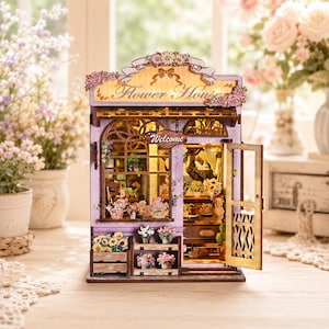 May include: A miniature wooden flower house model featuring a lavender exterior, a large front window, and an open door. The sign above the entrance reads Flower House, with a small hanging sign that says Welcome. The interior is illuminated.