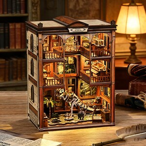 Op de afbeelding: Gedetailleerd houten model van een museum met meerdere niveaus, miniatuurexposities en trappen. Het model bevat een dinosaurus skelet, schilderijen en de tekst "Fossil Museum". De binnenkant is verlicht, wat een warme gloed cre&euml;ert.