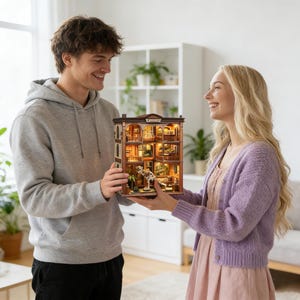 Op de afbeelding: Een gedetailleerd miniatuurpoppenhuis met binnenverlichting. De meerlaagse structuur heeft een bruine gevel met architectonische details. Het interieur is gemeubileerd en bevat een klein hondenbeeld. Het poppenhuis wordt door twee personen vastgehouden.