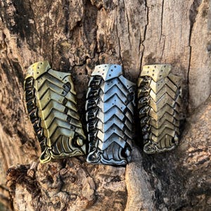 Könnte beinhalten: Drei metallische Miniatur-Rüstungsteile in Gold-, Silber- und Bronzetönen. Jedes Teil hat ein geschichtetes, schuppenartiges Design mit Nieten und einer gebogenen Basis. Die Teile werden auf einer strukturierten, braunen Holzoberfläche präsentiert.