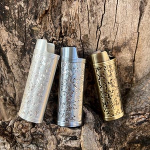 May include: Three metal lighters with ornate floral patterns. One is silver, one is a lighter shade of silver, and the third is gold. The lighters are standing upright against a textured, brown background.