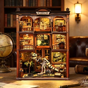 Op de afbeelding: Een gedetailleerd houten model van een fossielenmuseum, met meerdere niveaus met miniatuurexposities van dinoskeletten, fossielen en diorama's. Het model heeft een warme, uitnodigende gloed van binnenverlichting, met de tekst "Fossil Museum" op de gevel.