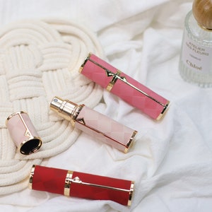 Puede incluir: Varios atomizadores de perfume en tonos rosa y rojo, con detalles dorados. Los atomizadores tienen un diseño geométrico y están colocados sobre una tela blanca. También hay un frasco de perfume transparente con tapón de madera.