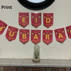 Może przedstawiać: Świąteczny baner z napisem "EID MUBARAK" w żółtych literach na czerwonych proporczykach. Każdy proporczyk zdobi wzór latarni. Baner wisi na białej ścianie, tworząc świąteczną atmosferę.