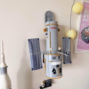 May include: A grey and gold brick-built model of the Hubble Space Telescope featuring blue solar panels, a hinged aperture door, and the NASA logo printed on the main cylindrical body. A small black display stand with a plaque sits below the model.