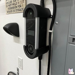 May include: A black electric vehicle charging station with a digital display and a black cord. The charging station is mounted on a white wall.