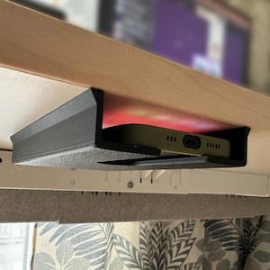 May include: A black plastic phone holder mounted beneath a wooden desk. The holder is designed to hold a smartphone securely in place.