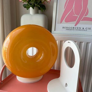 May include: An orange glass donut-shaped lamp with a white base sits on a pink table. A white, curved wall shelf is next to the lamp. A white vase with pink roses and a framed print with text are in the background.