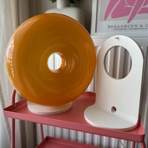 Pu&ograve; includere: Una lampada rotonda in vetro arancione con un foro centrale, appoggiata su una base di legno. Accanto, una mensola bianca ad arco. Entrambi sono su un carrello rosa in metallo.