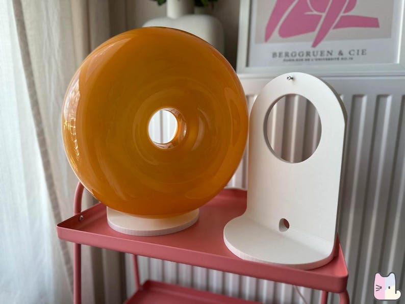 Pu&ograve; includere: Una lampada rotonda in vetro arancione con un foro centrale, appoggiata su una base di legno. Accanto, una mensola bianca ad arco. Entrambi sono su un carrello rosa in metallo.