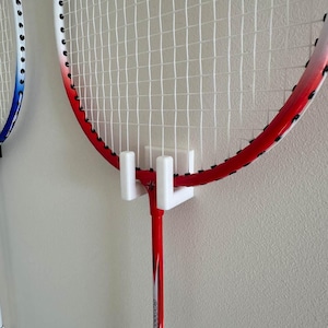 May include: A white wall mount with two white hooks holds a red and white badminton racket. The racket has a black grip and a white stringed head.