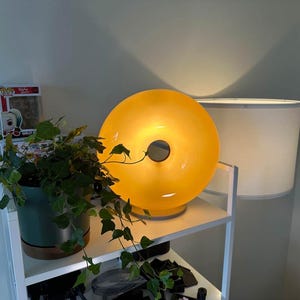 Pu&ograve; includere: Una moderna lampada illuminata a forma di ciambella giallo-arancio con un'apertura circolare al centro. La lampada &egrave; appoggiata su una mensola bianca, accanto a un paralume bianco e a una pianta in vaso con foglie verdi. &Egrave; visibile una statuetta Funko Pop.