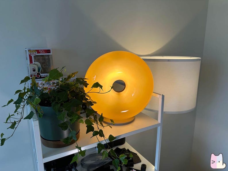 Pu&ograve; includere: Una moderna lampada illuminata a forma di ciambella giallo-arancio con un'apertura circolare al centro. La lampada &egrave; appoggiata su una mensola bianca, accanto a un paralume bianco e a una pianta in vaso con foglie verdi. &Egrave; visibile una statuetta Funko Pop.