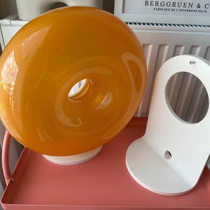 Pu&ograve; includere: Una lampada in vetro arancione con un design circolare e una base bianca. Accanto, una mensola da parete bianca e curva con un'apertura circolare. Gli oggetti sono su un vassoio rosa.