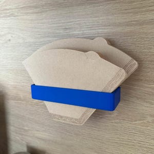 Peut inclure: Deux filtres à café en papier brun sont maintenus en place par un support en plastique bleu monté sur une surface en bois.