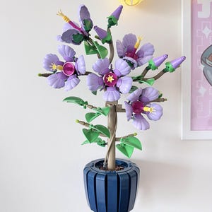 Pode incluir: Uma planta decorativa feita de blocos de plástico, com flores lavanda e estames amarelos. As folhas são verdes. A planta está num vaso azul escuro, fixado na parede. As flores têm um design detalhado, o vaso tem linhas verticais.