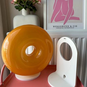 Pu&ograve; includere: Una lampada arancione a forma di ciambella in vetro &egrave; appoggiata su una base bianca, accanto a una mensola in legno bianca. Un vaso di rose rosa e una stampa incorniciata con figure rosa e la scritta "BERGGRUEN & CIE" sono sullo sfondo.