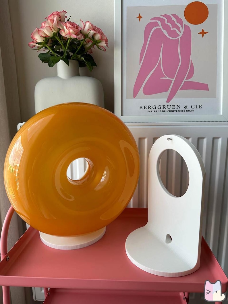 Pu&ograve; includere: Una lampada arancione a forma di ciambella in vetro &egrave; appoggiata su una base bianca, accanto a una mensola in legno bianca. Un vaso di rose rosa e una stampa incorniciata con figure rosa e la scritta "BERGGRUEN & CIE" sono sullo sfondo.