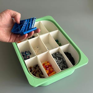 Könnte beinhalten: Eine hellgrüne Aufbewahrungsbox mit mehreren Fächern, gefüllt mit verschiedenen kleinen Bausteinen in verschiedenen Farben. Eine Hand hält ein blaues Bausteinteil über der Box.