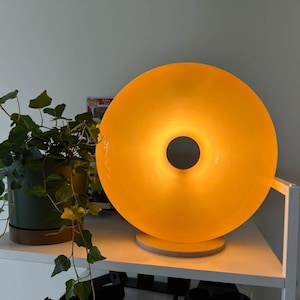 Pu&ograve; includere: Una moderna lampada gialla a forma di ciambella con un'apertura circolare al centro. La lampada poggia su una base rotonda ed emette un bagliore caldo. A sinistra della lampada c'&egrave; una pianta in vaso con foglie verdi.