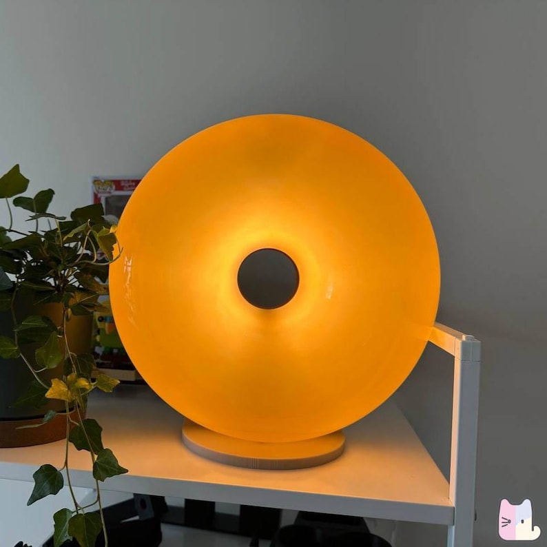 Pu&ograve; includere: Una moderna lampada da tavolo illuminata con un design circolare a forma di ciambella. La lampada emette una calda luce gialla e poggia su una base rotonda in legno. Un pezzo decorativo unico per un soggiorno o una camera da letto.