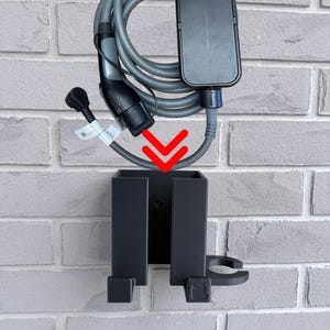 May include: A black EV charger holder mounted on a brick wall. The holder is designed to store the charging cable and connector. The charging cable is gray and coiled above the holder. The connector is black and gray.