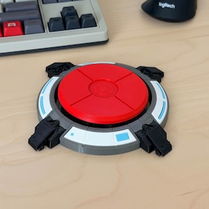 Könnte beinhalten: Ein 3D-gedrucktes Gerät mit roter kreisförmiger Oberseite und grauem Sockel. Der Sockel hat ein weiß-blaues Ringdesign und vier schwarze rechteckige Vorsprünge. Das Gerät befindet sich auf einer hellen Holzoberfläche, mit Tastatur und Maus im Hintergrund.