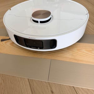 May include: A white circular robot vacuum cleaner featuring a metallic silver center sensor dome, a top-mounted control panel, and a front-facing camera module labeled with the text AI Action, designed for automated floor cleaning and navigation.