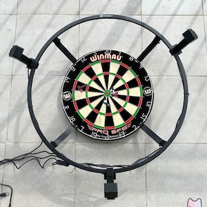 Op de afbeelding: Een zwarte dartbord met een rood en groen bullseye en een zwarte ring rond de buitenkant. Het dartbord is gemonteerd op een zwarte metalen standaard met een cirkelvormig frame. Het dartbord is gelabeld "Winmau" en "Pro SFB".