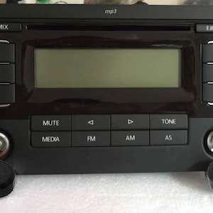 Può includere: Un'autoradio nera con un display digitale, pulsanti per i media, FM, AM e AS e un pulsante di silenziamento. L'autoradio ha una cornice argentata attorno ai bordi e una manopola argentata sul lato destro. L'autoradio è montata su un cruscotto di un'auto.