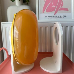 Pu&ograve; includere: Un vaso di vetro arancione con un accento giallo, posto su un supporto bianco. Accanto, un fermalibri bianco. Il vaso &egrave; su un carrello rosa, con una stampa incorniciata e un vaso bianco con fiori sullo sfondo.