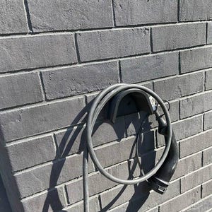 Pode incluir: Um cabo elétrico preto está enrolado e preso a uma parede de tijolo com um suporte de plástico preto.