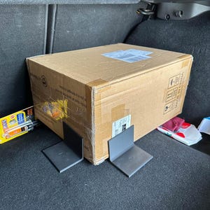 Könnte beinhalten: Ein großer brauner Karton, mit Klebeband versiegelt, befindet sich im Kofferraum eines Fahrzeugs. Zwei dunkelgraue Metall-L-Winkel befinden sich vor der Box. Weitere Gegenstände sind im Hintergrund sichtbar.