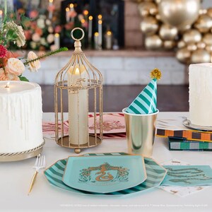 Puede incluir: Una mesa festiva con un candelabro dorado en forma de jaula, platos turquesa con un escudo y un gorro de fiesta. La escena incluye una copa dorada, velas y arreglos florales, creando un ambiente festivo.