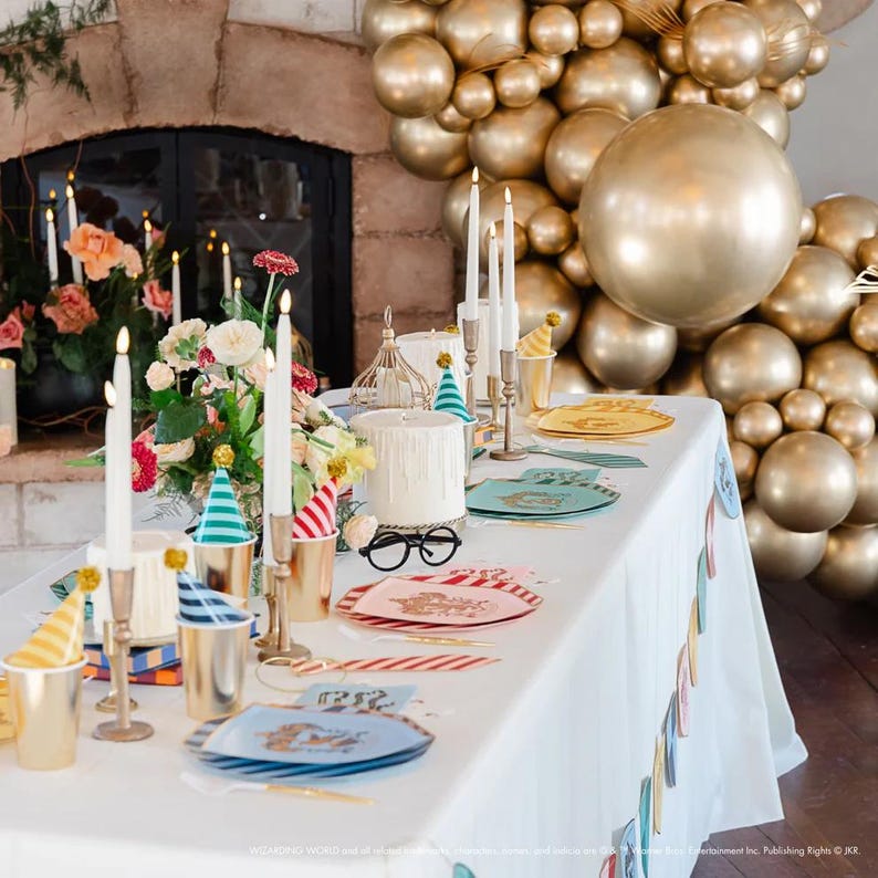 Puede incluir: Una mesa de fiesta decorada con un mantel blanco, globos dorados y velas. La mesa est&aacute; decorada con gorros de fiesta, platos y copas doradas. Tambi&eacute;n hay un pastel y un arreglo floral, creando un ambiente festivo.