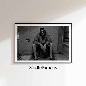 May include: Black and white photograph of a man sitting on a toilet in a bathroom. He is wearing a robe and has a beard. The bathroom has patterned wallpaper and a window with a curtain.