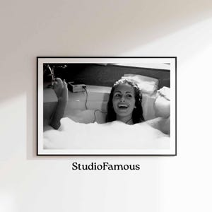 May include: Black and white photograph of a woman in a bath, smiling and listening to music. She is wearing a white towel and has her hair pulled back. The bath is filled with bubbles.