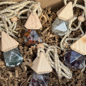 Könnte beinhalten: Fünf kleine Glasflaschen mit verschiedenen farbigen Kristallen, jede Flasche ist an einem dreieckigen Holzanhänger und einer goldenen Schnur befestigt.