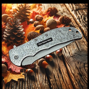 May include: A silver pocket knife with an engraved design and the text "Your Name Here" on the handle. The knife is lying on a wooden surface with autumn leaves and acorns.